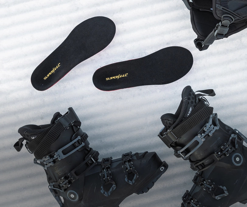 Black ski boots laid on some snow with Superfeet's Winter Think Support Insole laying near by. 