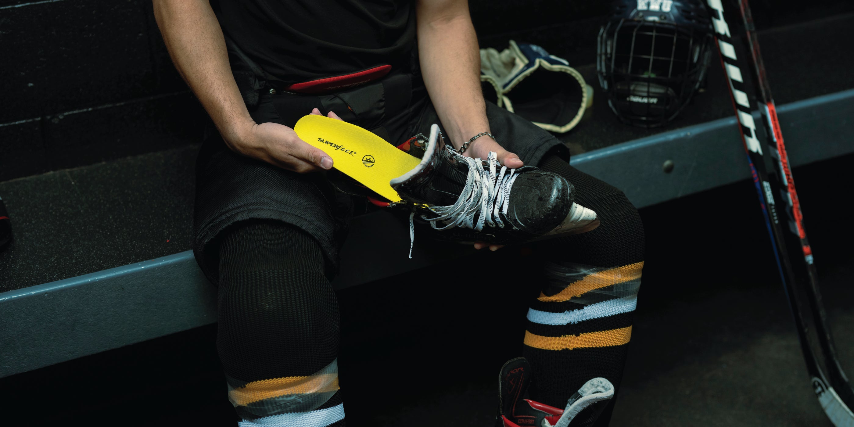 Hockey player sitting on bench in ice arena inserting a Superfeet Hockey Performance Insole into a hockey ice skate.
